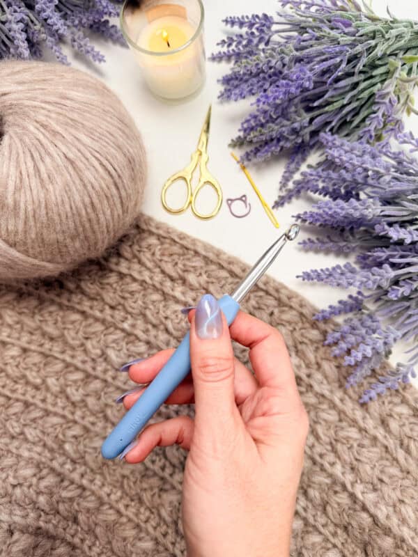 A hand holding a blue crochet hook above a textured brown yarn project, with a ball of yarn, gold scissors, stitch markers, a candle, and lavender flowers nearby.