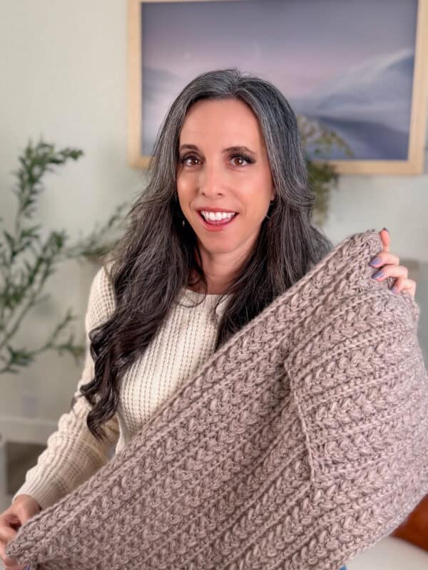 A woman with long gray hair holds up a chunky knitted beige blanket, smiling indoors with plants and a framed picture in the background.