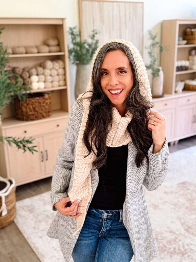 Woman smiling indoors, wearing a gray coat, black top, jeans, and a cream knit scarf with hood crafted using the Wheat Stitch™ crochet hooded scarf pattern. Wooden shelves with baskets and yarn are in the background.