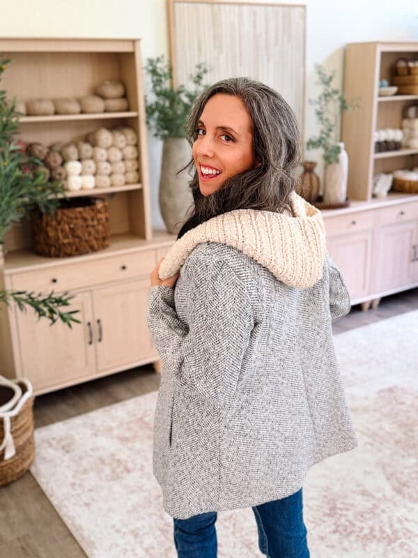 Woman with long brown hair and gray jacket turns and smiles in a cozy, well-decorated living room with shelves and yarn in the background, perhaps inspired by crafting her own Wheat Stitch™ crochet hooded scarf pattern.