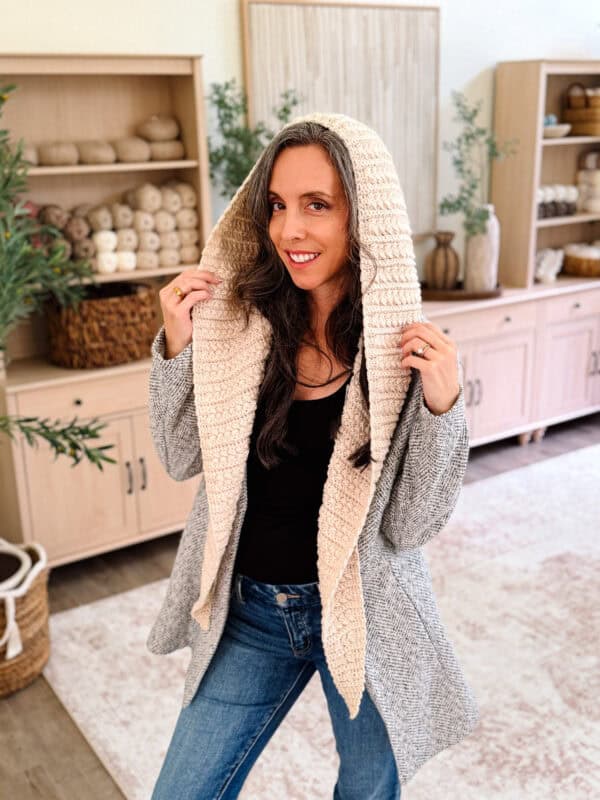 Woman indoors wearing a gray cardigan with a large, handcrafted hood made using the Wheat Stitch™ crochet hooded scarf pattern, standing in a cozy room with shelves, yarn, and decor in the background.