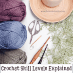 Four skeins of yarn, a candle, crochet hooks, and scissors arranged on a white surface with the text: "Crochet Skill Levels Explained. Discover crochet skill levels—no pressure, just helpful guidance.