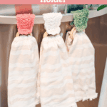 Three white striped dishtowels hang from a bar with crocheted holders in pink, white, and green. The image promotes a free Wheat Stitch napkin ring crochet pattern and video tutorial.