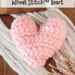 A pink crocheted heart made with wheat stitch is displayed on a wooden surface with text advertising a free pattern and video tutorial.