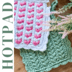 Two crocheted hot pads, one white and pink with a scalloped edge and one green, displayed on a wooden surface with decorative foliage. Text reads "Wheat Stitch™ HOTPAD.