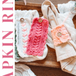 Three crocheted napkin rings in shades of pink and cream, each with a button, displayed on and beside fabric napkins. Inspired by the Wheat Stitch mug cozy crochet pattern. Text reads "Napkin Ring Wheat Stitch.