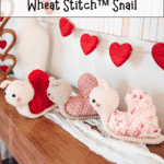 Three crocheted snail plushies in shades of red, pink, and cream are displayed on a shelf beneath a decorative garland of red hearts made with the Wheat Stitch crochet heart pattern.
