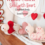 Three crocheted snail plushies with heart-shaped shells, made using the Wheat Stitch crochet heart pattern in red, pink, and white, are displayed on a wooden surface, with heart decorations hanging in the background.