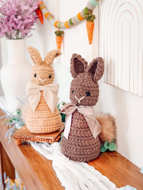 Two crocheted bunny figures with bows are displayed on a wooden shelf, with a felt carrot garland and vase of flowers in the background.