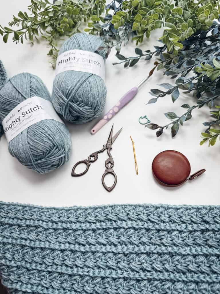 Two skeins of blue yarn, crochet hooks, scissors, a needle, measuring tape, and a partially crocheted piece are arranged on a white surface with some green leaves.