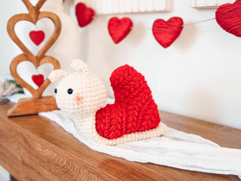 A crocheted snail with a red, Wheat Stitch crochet heart pattern shell sits on a white cloth atop a wooden surface, with red heart decorations in the background.