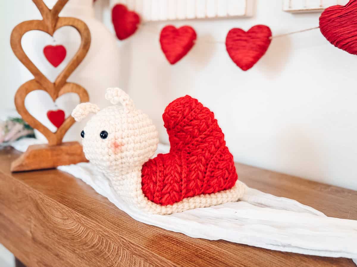 A crocheted snail with a red, Wheat Stitch crochet heart pattern shell sits on a white cloth atop a wooden surface, with red heart decorations in the background.