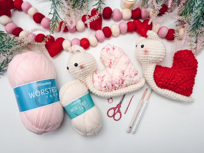Two skeins of worsted yarn, crochet hooks, scissors, and two crocheted snail toys are arranged on a white surface with decorative garlands and greenery.