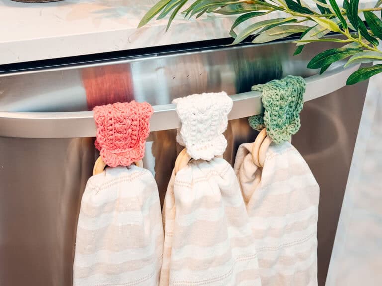 Three striped dish towels with knitted tops in pink, white, and green hang on a stainless steel appliance handle, accented by a Wheat Stitch napkin ring crochet pattern. A leafy branch peeks in on the right.