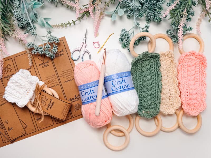 Crochet kit with yarn, pattern cards, crochet hook, scissors, stitch marker, finished Wheat Stitch napkin ring crochet pattern pieces with wooden rings, and sprigs of artificial greenery arranged on a white surface.