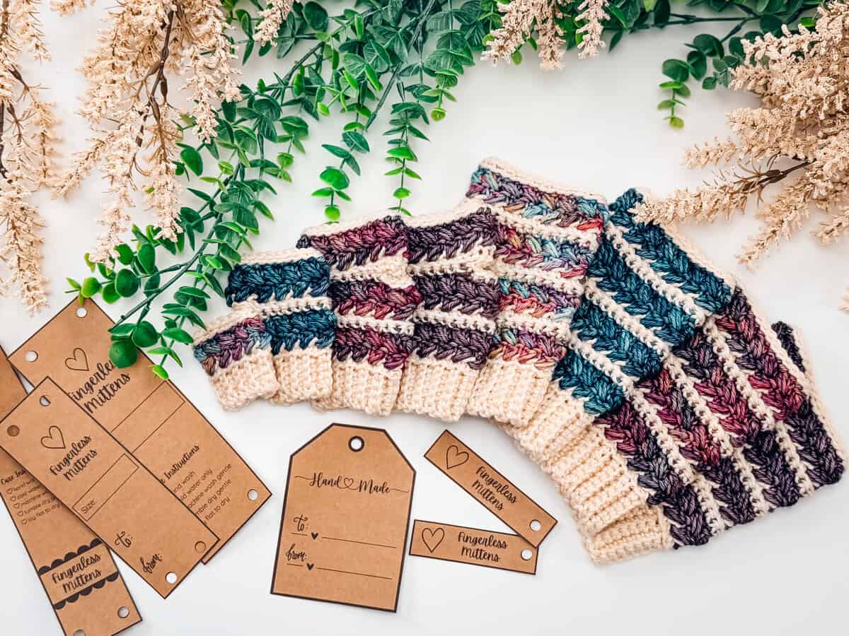 Crocheted mittens with striped patterns in cream, purple, and teal yarn are displayed on a white surface with brown product tags and green foliage accents.