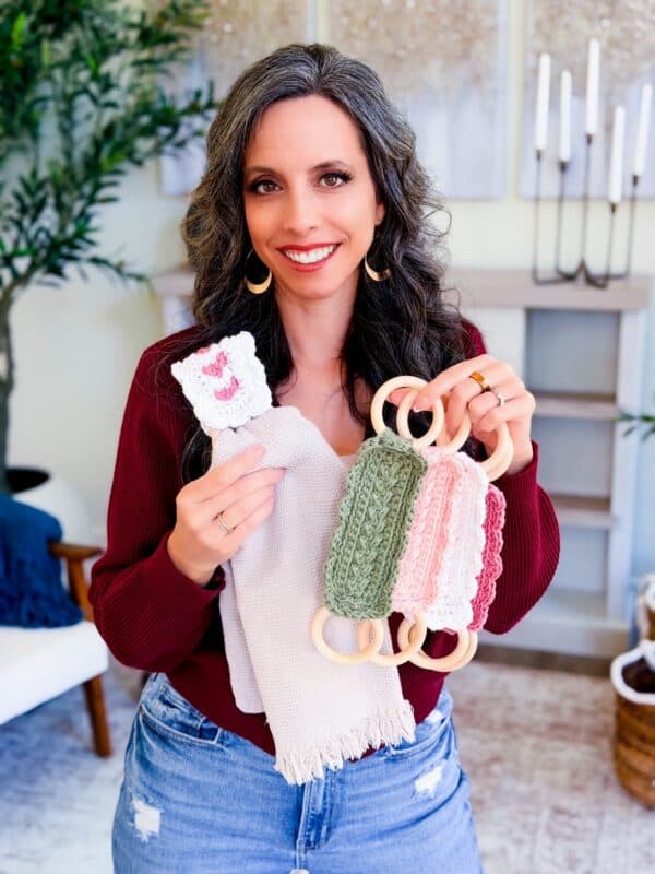 A woman with long, wavy gray hair holds crocheted items with wooden rings and a small white cloth with a pink heart, displaying a Wheat Stitch napkin ring crochet pattern while standing in her living room.