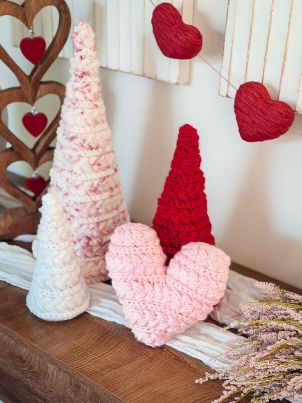 Three crocheted cones and a pink crocheted heart sit on a wooden surface, with red heart decorations and a garland hanging in the background.