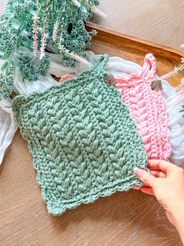 A hand holds a chunky green crochet square with a scalloped edge, next to a pink crochet square, on a wooden tray with decorative greenery and fabric.