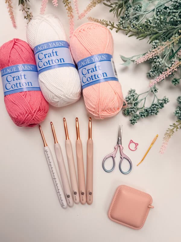 Three skeins of craft cotton yarn, crochet hooks, scissors, a stitch marker, a yarn needle, and a pink case are arranged on a white surface with decorative foliage.