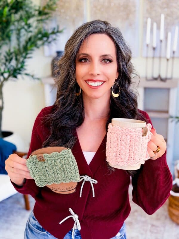Woman with long brown and gray hair, wearing a maroon sweater, smiles at the camera while holding two mugs wrapped in Wheat Stitch mug cozy crochet pattern covers&mdash;one green, one pink&mdash;in a cozy indoor setting.