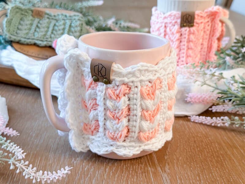 A light pink mug with a crocheted white and peach cozy, inspired by the Wheat Stitch hotpad crochet pattern, sits on a wooden surface surrounded by similar mugs and decorative faux greenery.