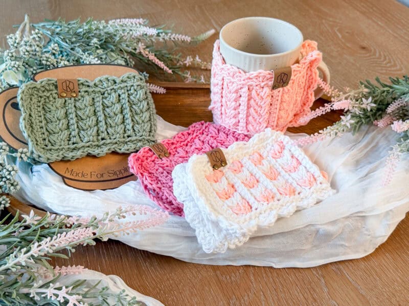 Four hand-knitted mug cozies in pastel colors are displayed on a wooden table with a mug, flowers, cloth, and a Wheat Stitch hotpad crochet pattern, creating a cozy and decorative arrangement.
