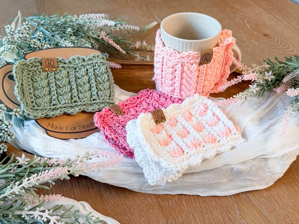 Four hand-knitted mug cozies in pastel colors are displayed on a wooden table with a mug, flowers, cloth, and a Wheat Stitch hotpad crochet pattern, creating a cozy and decorative arrangement.