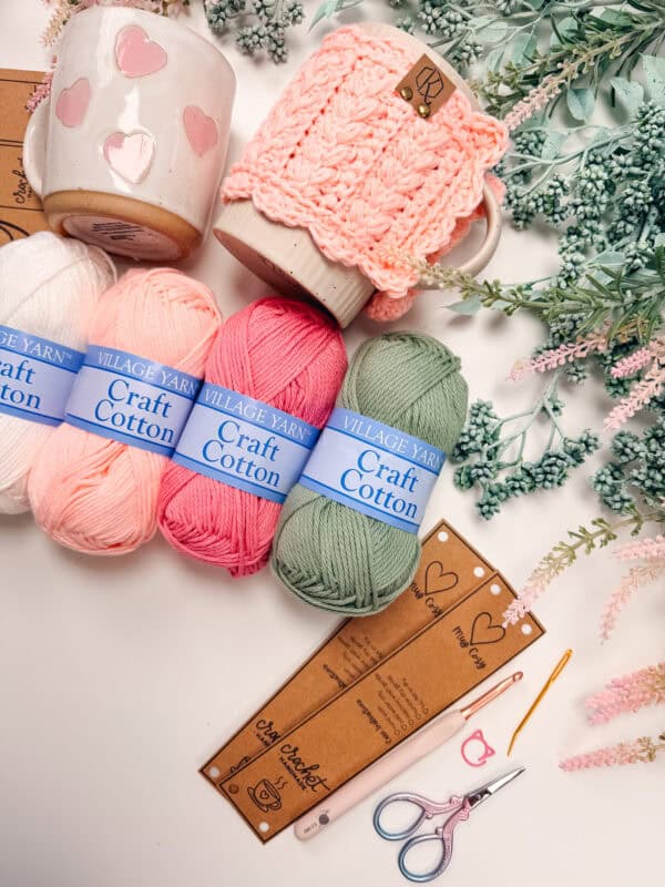 Three yarn-wrapped mugs, skeins of cotton yarn, crochet tools, scissors, and sprigs of faux greenery arranged on a white surface&mdash;perfect inspiration for your next Wheat Stitch hotpad crochet pattern project.