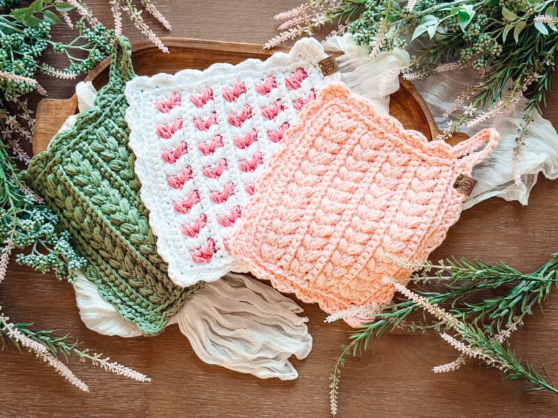 Three crocheted potholders in green, pink, and peach are displayed on a wooden tray surrounded by greenery and white fabric.