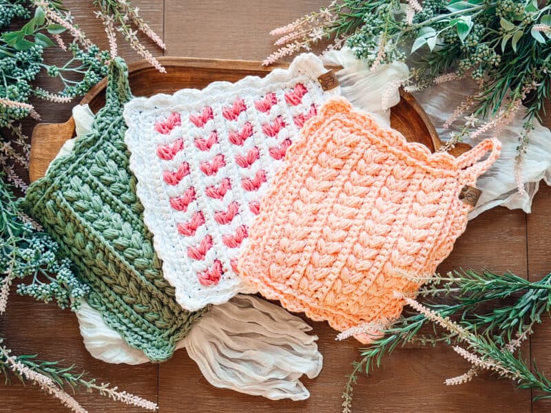 Three crocheted pot holders in green, white with pink, and peach colors are arranged on a wooden tray with greenery and fabric accents around them.