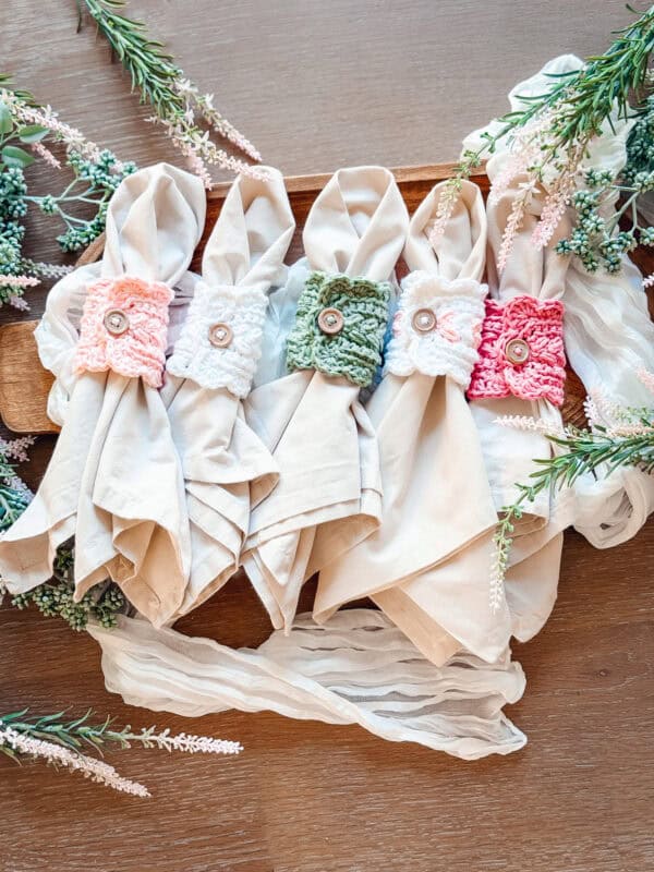Five beige cloth napkins with crocheted napkin rings—reminiscent of the Wheat Stitch mug cozy crochet pattern—in pastel colors, each with a button in the center, arranged on a wooden tray with greenery and fabric accents.