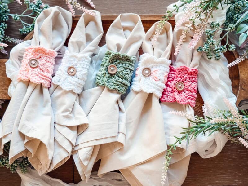 Four beige napkins are each held by a different colored crocheted napkin ring with a button, arranged on a wooden tray and surrounded by greenery&mdash;perfectly complementing the Wheat Stitch mug cozy crochet pattern.