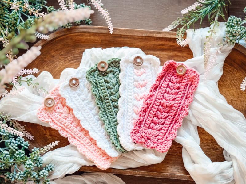 Five crocheted bookmarks in pink, white, green, and peach tones with button closures are displayed on a wooden tray surrounded by greenery and white fabric, pairing beautifully with a Wheat Stitch mug cozy crochet pattern.