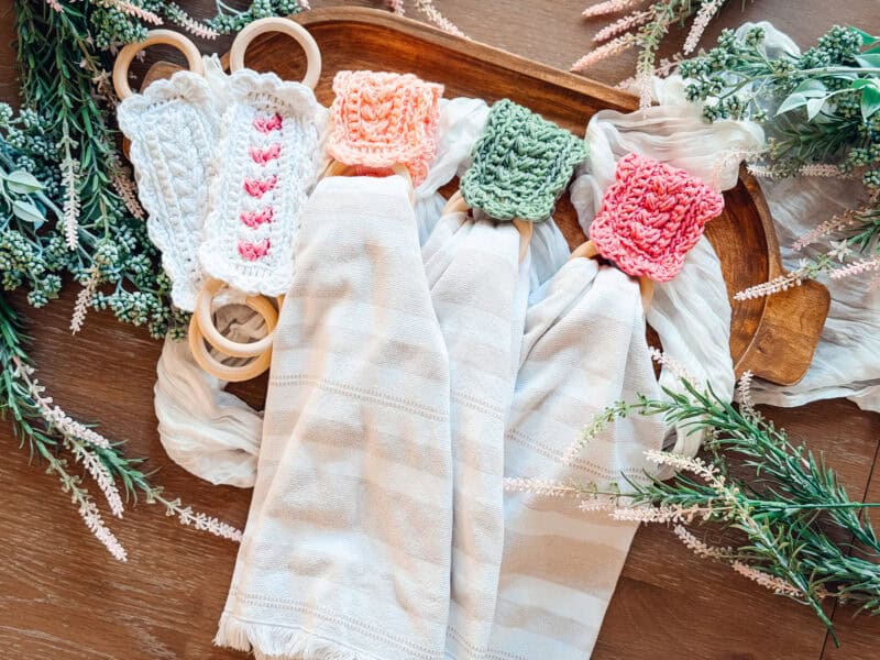A wooden tray holds three striped cloth napkins with Wheat Stitch napkin ring crochet patterns in orange, pink, and green, surrounded by greenery and a pair of white crocheted baby rattles.