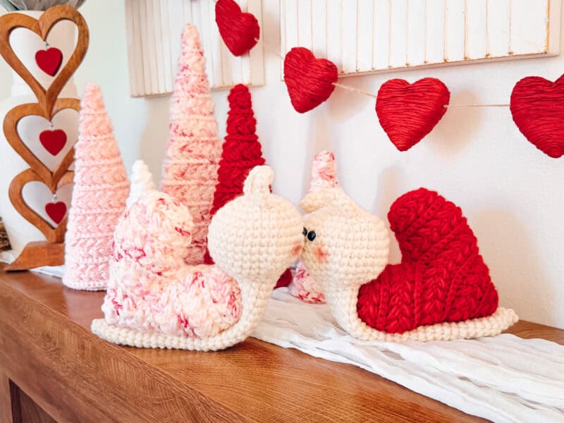 Two crocheted snails with heart-shaped shells, crafted using the Wheat Stitch crochet heart pattern, face each other on a wooden shelf, surrounded by pink and red yarn trees and heart decorations.