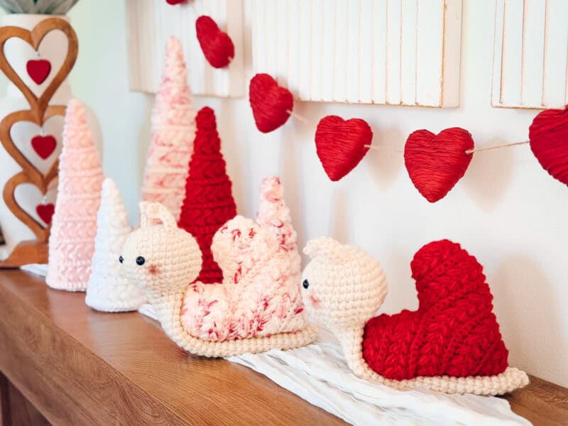 Crocheted snail and tree decorations in red, white, and pink sit on a wooden surface, while a Wheat Stitch crochet heart pattern garland hangs above.