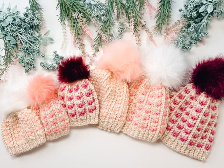 Five crocheted hats with pink and burgundy accents and large pom-poms are arranged in a row on a white surface with greenery and pink floral decorations above them.