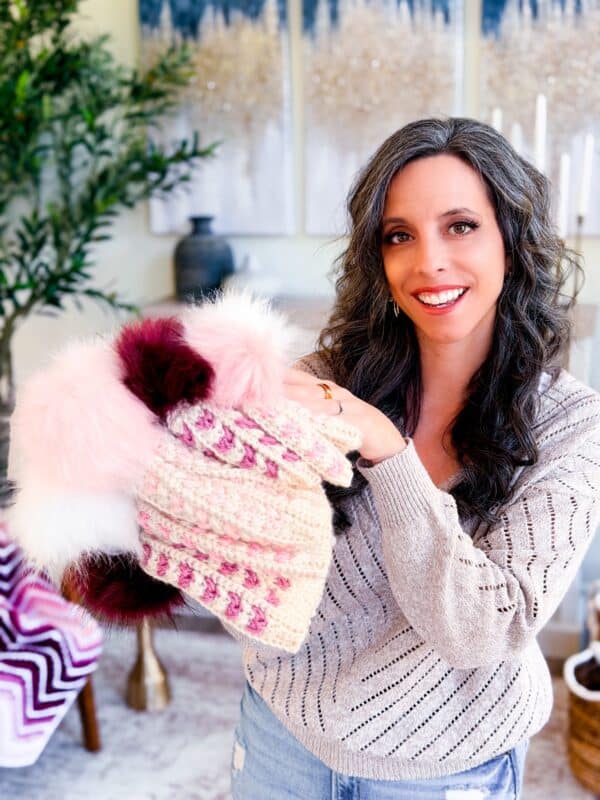 A woman with long, wavy hair smiles while holding several knitted hats with pom-poms in a cozy, decorated living room.