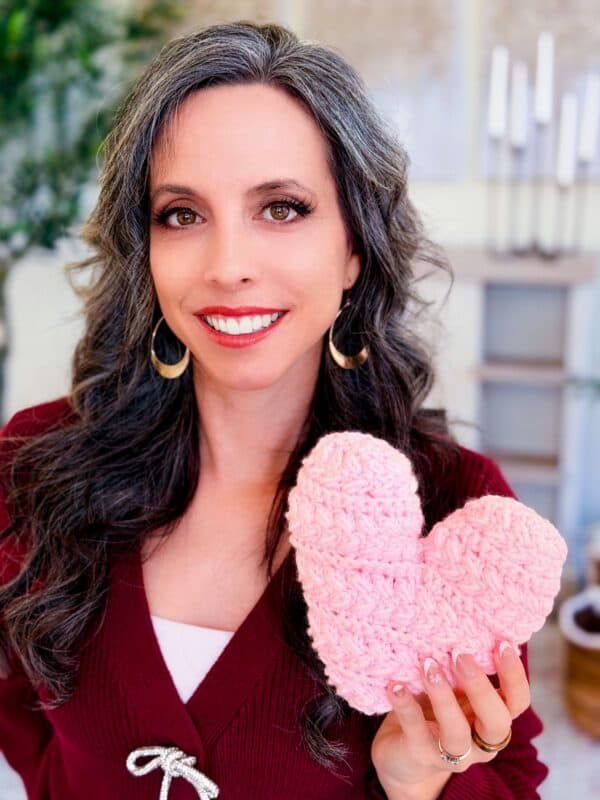 Woman with long, wavy gray hair and a burgundy top smiles while holding a pink, crocheted heart-shaped object in her hand.