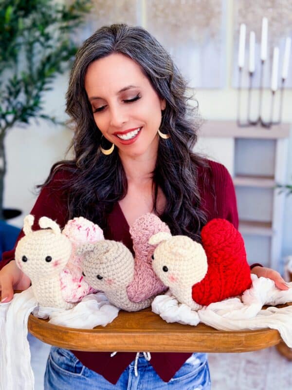 A woman with long dark hair holds a wooden tray displaying four crocheted snail toys in cream, pink, and red—each featuring details inspired by the Wheat Stitch crochet heart pattern.