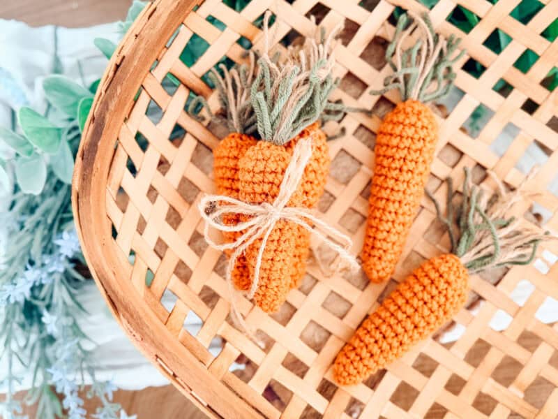 Three crocheted carrots with green tops are arranged in a woven basket&mdash;two tied together with simple string. Pair them with a Crochet Bunny Head Pattern for an adorable springtime display.