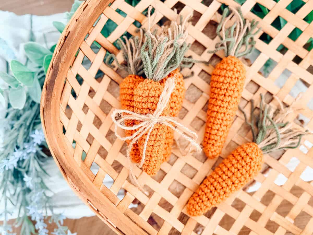Three crocheted carrots with green tops are arranged in a woven basket—two tied together with simple string. Pair them with a Crochet Bunny Head Pattern for an adorable springtime display.