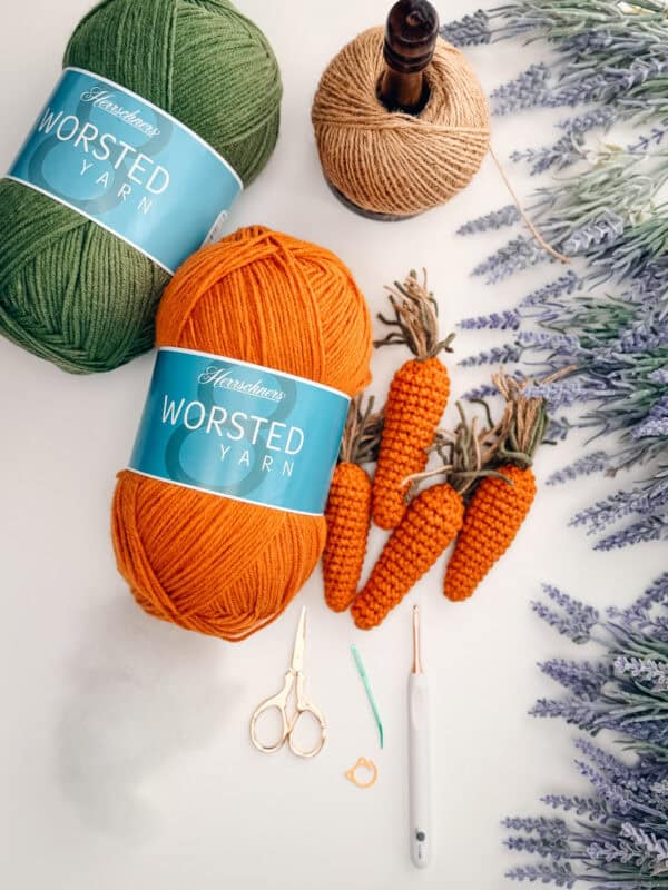 Two skeins of green and orange worsted yarn, a spool of tan thread, crocheted carrots, scissors, a crochet hook, a needle, artificial lavender, and a Crochet Bunny Head Pattern are arranged on a white surface.