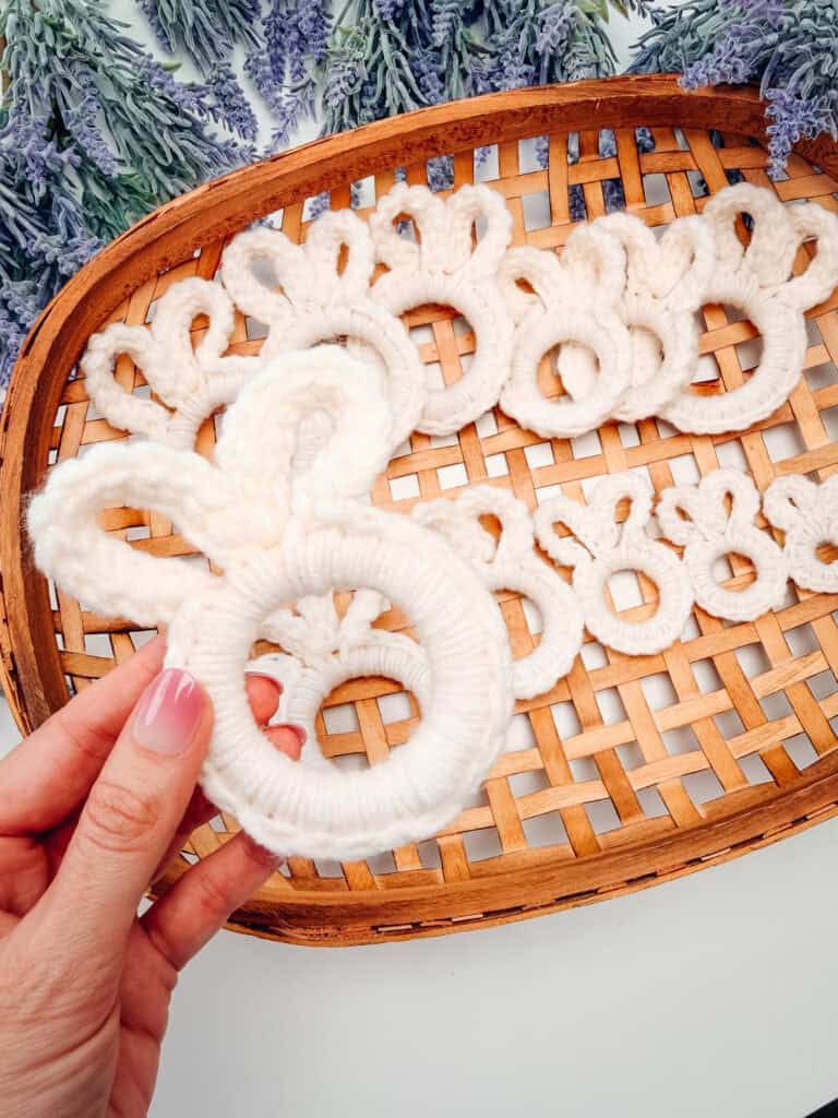 A hand holding a white yarn ring shaped like a bunny on a woven tray with similar bunny shapes; lavender flowers are in the background.