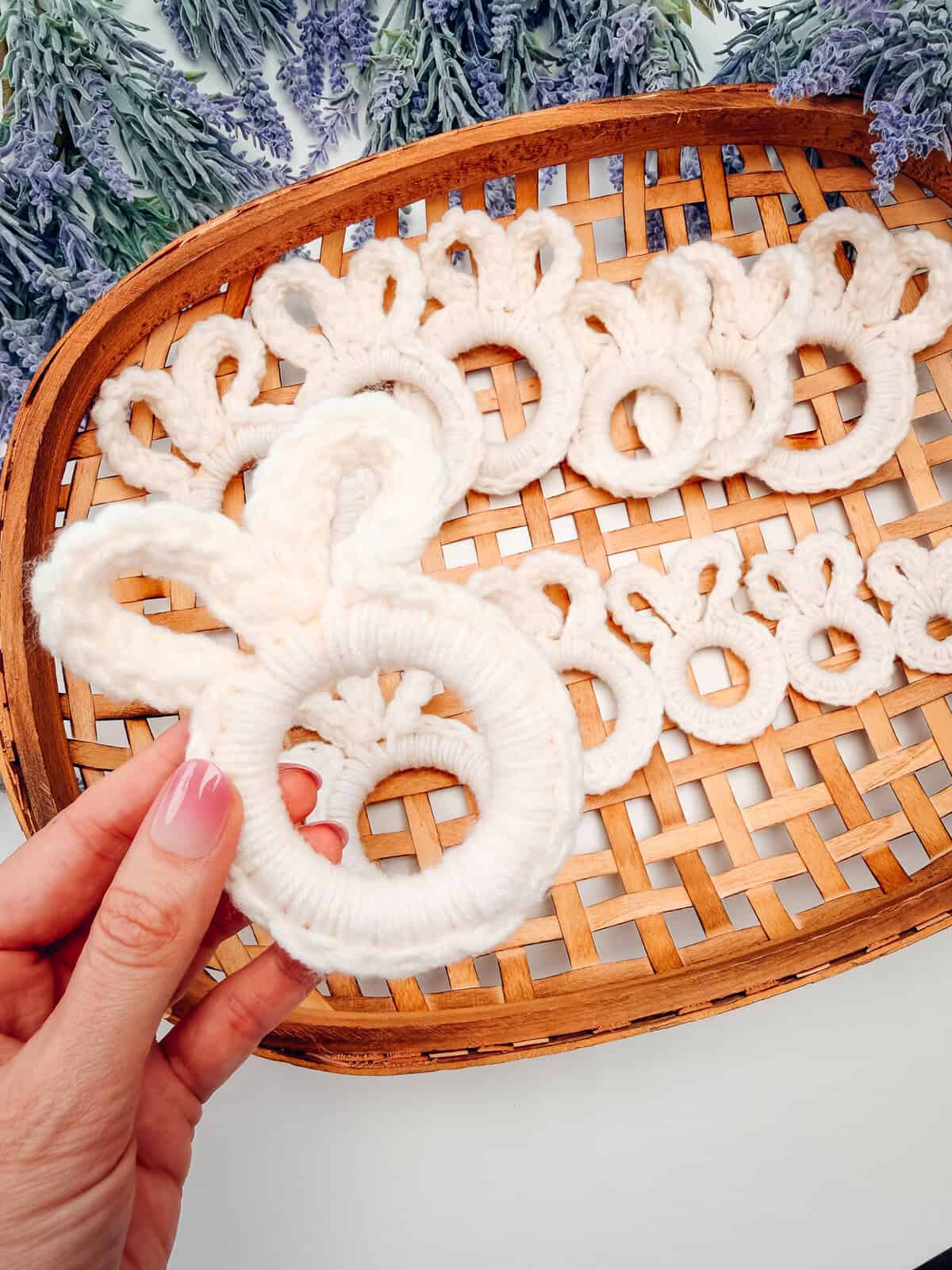 A hand holding a white yarn ring shaped like a bunny on a woven tray with similar bunny shapes; lavender flowers are in the background.