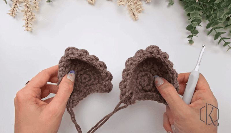 Two hands hold two brown crocheted pieces with scalloped edges, crafted using the Wheat Stitch Bunny Crochet Pattern; a crochet hook lies nearby. Greenery and dried flowers provide a charming backdrop.