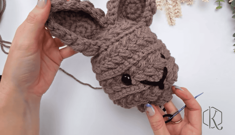 A person holds a partially crocheted brown bunny head made with the Wheat Stitch Bunny Crochet Pattern, featuring black safety eyes and a yarn needle threaded with black yarn for sewing facial features.