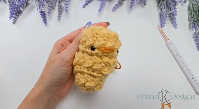 A hand holding a crocheted yellow wheat stitch chick with black eyes and an orange beak; a crochet hook and purple flowers are nearby.