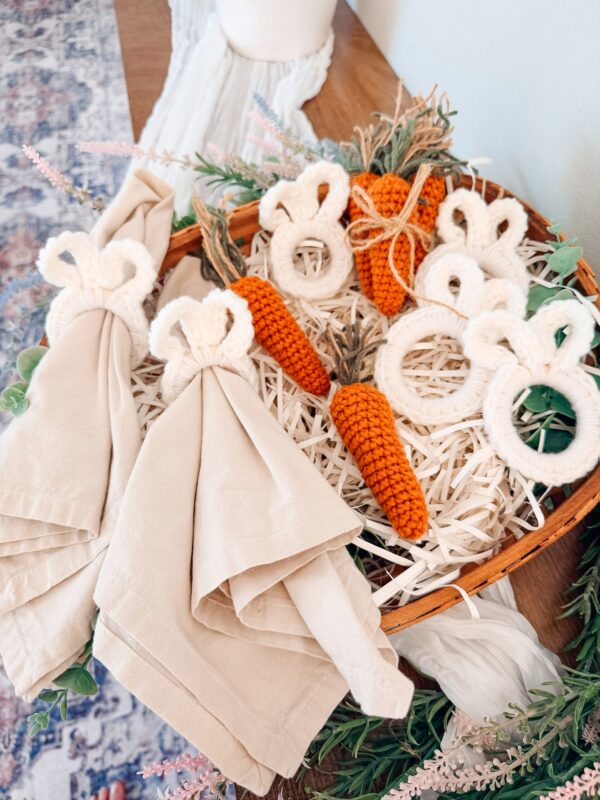 A basket holds beige napkins with crocheted bunny napkin rings and a few carrots made from the Mini Crochet Carrot Pattern, surrounded by decorative greenery.